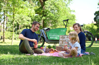 Eine glückliche Familie sitz beim Picknick im Park. Im Hintergrund steht ein Lastenrad mit dem Aufdruck: Sigo E-Lastenrad Sharing.
